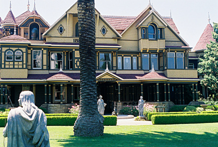 [A long three-story yellow building with brown trim. There are many gables and points and other shapes to the roofline. Many windows across the front of many different shapes and sizes. A squared-off hedge is in front of the house and there are numerous statues in the yard.]